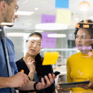 Three individuals gather around a table, discussing ideas with sticky notes displayed on the wall behind them.