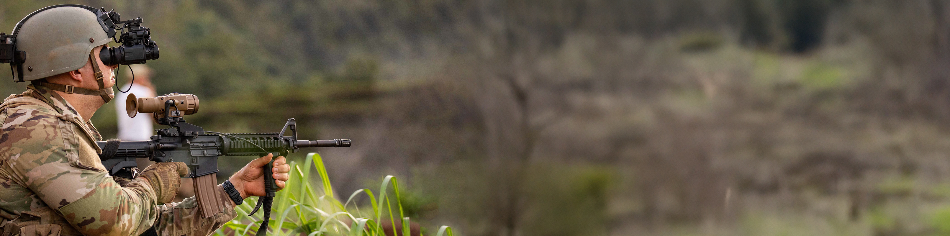A soldier aims a firearm at a target hidden in the grass, focused and alert in a tactical stance.
