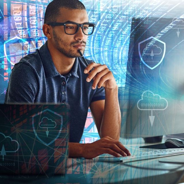 A man wearing glasses is seated at a desk, working on a laptop with a cloud icon displayed nearby.